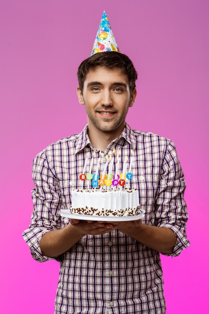 Giovane uomo bello che sorride, tenendo la torta di compleanno sopra la parete viola.