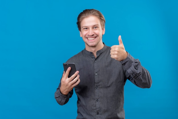 Giovane uomo bello che tiene il telefono cellulare che mostra i pollici in su sorridente in piedi sopra la parete blu