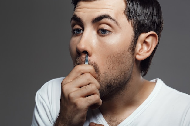 Giovane uomo bello che tira capelli nel naso sopra la parete grigia