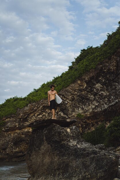 Giovane uomo bello con una tavola da surf su una roccia vicino all'oceano.