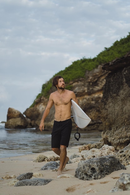 Giovane uomo bello con una tavola da surf su una roccia vicino all'oceano.