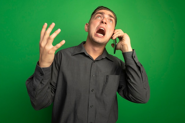 Giovane uomo bello frustrato in camicia grigia che grida e urla mentre parla sul telefono cellulare