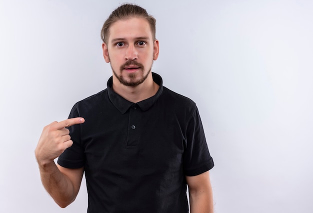 Giovane uomo bello in camicia di polo nera che sembra spaventato che punta a se stesso in piedi su sfondo bianco