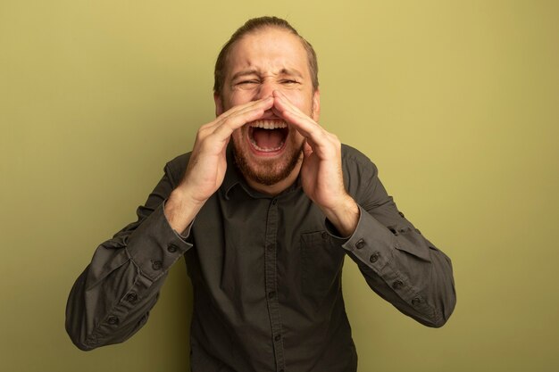 Giovane uomo bello in camicia grigia che grida o chiama qualcuno con le mani vicino alla bocca con espressione infastidita