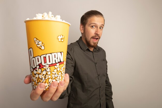 Giovane uomo bello in camicia grigia che mostra secchio con popcorn sorpreso