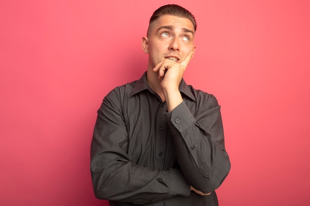 Giovane uomo bello in camicia grigia che osserva in su in piedi perplesso sopra il muro rosa