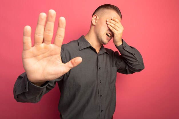 Giovane uomo bello in camicia grigia che sembra stanco e annoiato facendo gesto di arresto con la mano aperta in piedi sopra il muro rosa