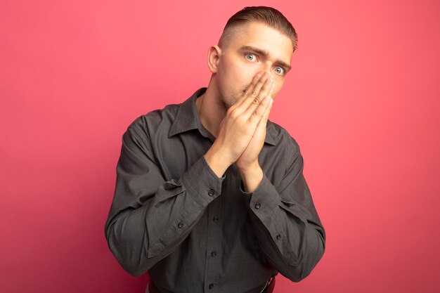 Giovane uomo bello in camicia grigia che tiene le palme insieme sul suo viso che sembra preoccupato in piedi sopra il muro rosa