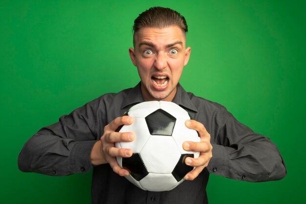 Giovane uomo bello in camicia grigia che tiene pallone da calcio gridando con espressione aggressiva