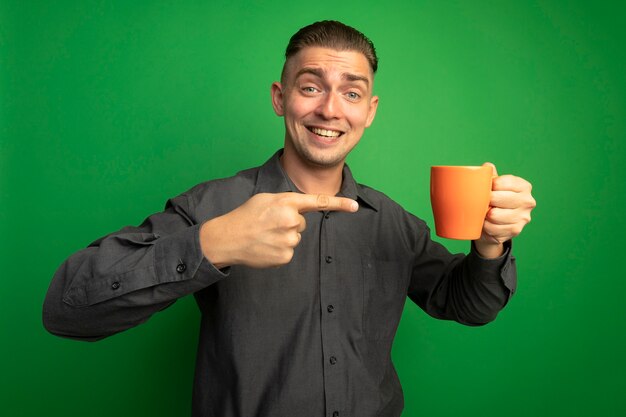 Giovane uomo bello in camicia grigia che tiene tazza arancione che punta con il dito indice sorridendo allegramente in piedi sopra la parete verde