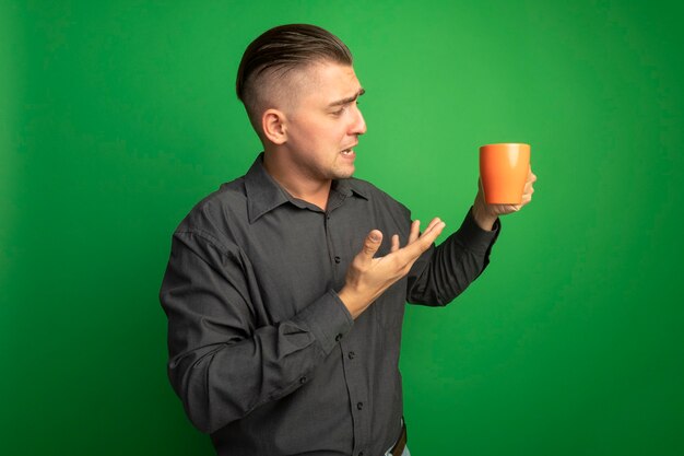 Giovane uomo bello in camicia grigia che tiene tazza arancione presentandolo con il braccio che è confuso