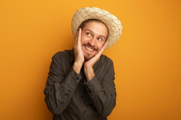 Giovane uomo bello in camicia grigia e cappello estivo che osserva da parte con le braccia sul viso felice e positivo