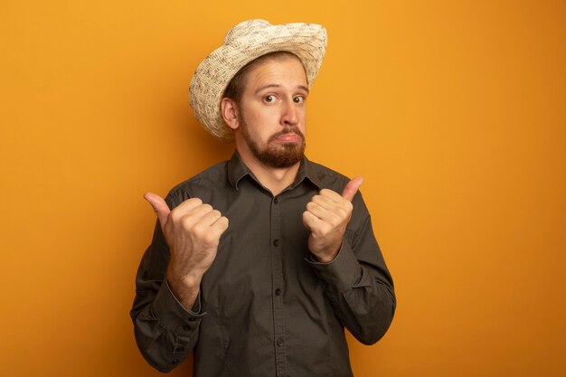 Giovane uomo bello in camicia grigia e cappello estivo lookign alla telecamera confuso che mostra i pollici in su