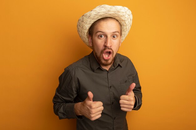 Giovane uomo bello in camicia grigia e cappello estivo sorpreso e felice che mostra i pollici in su