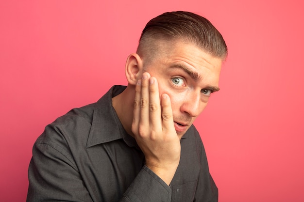 Giovane uomo bello in camicia grigia guardando la parte anteriore confusa con il braccio sulla guancia in piedi sopra il muro rosa