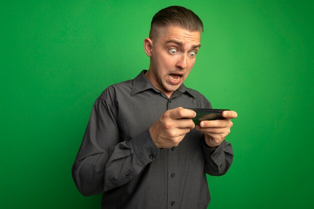Giovane uomo bello in camicia grigia utilizza lo smartphone essendo sorpreso e stupito in piedi sopra la parete verde