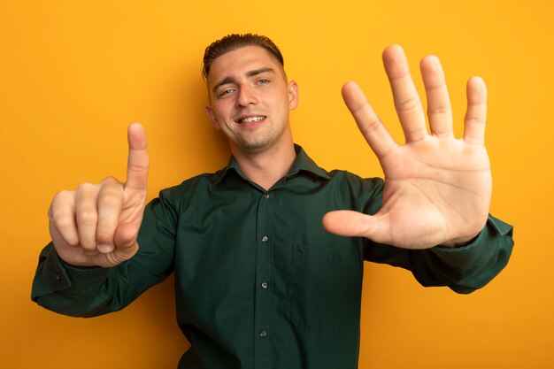 Giovane uomo bello in camicia verde che mostra e rivolto verso l'alto con il dito numero sei sorridendo allegramente