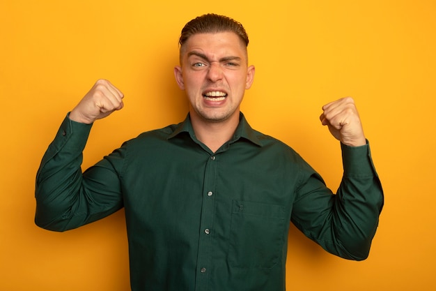 Giovane uomo bello in camicia verde che stringe i pugni felici ed eccitati che rallegrano il suo successo in piedi sopra la parete arancione