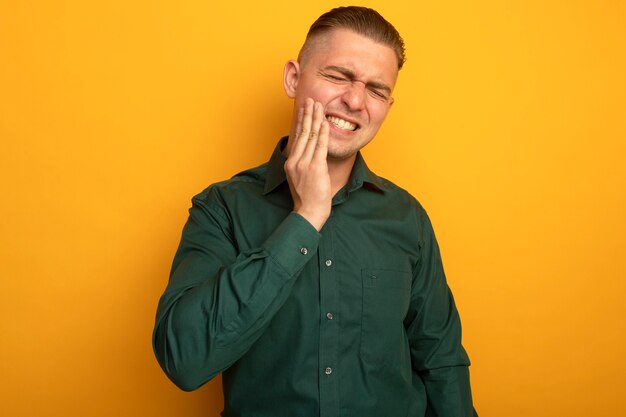 Giovane uomo bello in camicia verde che tocca la sua guancia che ha mal di denti