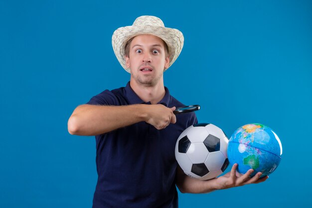 Giovane uomo bello in cappello estivo tenendo palla da calcio e globo andando a guardare il globo attraverso la lente di ingrandimento guardando sorpreso e stupito in piedi su sfondo blu