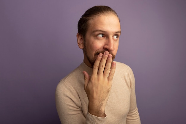 Giovane uomo bello in dolcevita beige che osserva da parte sorpreso che copre la bocca con la mano