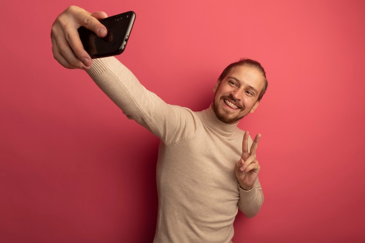 Foto gratuito giovane uomo bello in dolcevita beige utilizza lo smartphone facendo selfie sorridente che mostra v-segno