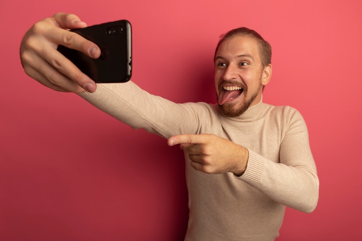 Foto gratuito giovane uomo bello in dolcevita beige utilizza lo smartphone facendo selfie sorridente spuntavano lingua in piedi sopra il muro rosa