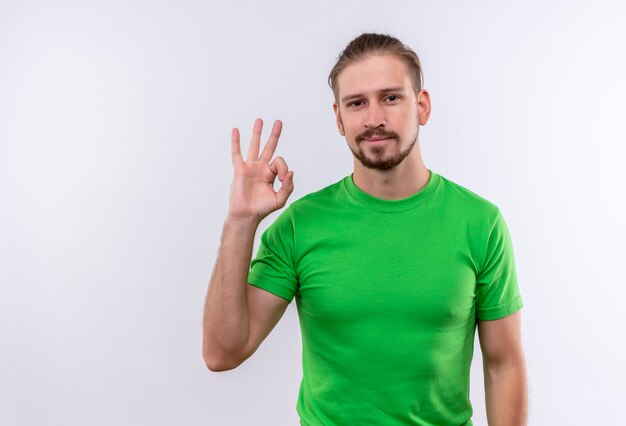 Giovane uomo bello in maglietta verde che guarda l'obbiettivo che sorride puramente facendo segno giusto in piedi su sfondo bianco