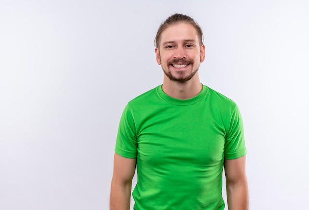 Giovane uomo bello in maglietta verde che guarda l'obbiettivo sorridente allegramente in piedi su sfondo bianco