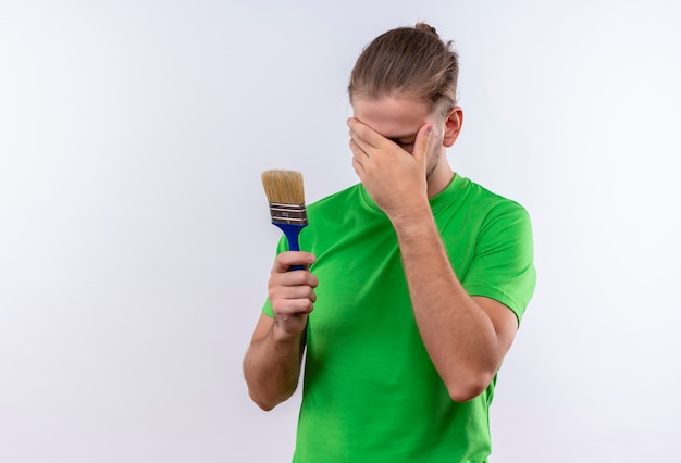 Giovane uomo bello in t-shirt verde che tiene il pennello che copre il viso con la mano in piedi su sfondo bianco