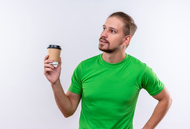 Giovane uomo bello in t-shirt verde che tiene tazza di caffè che osserva da parte con l'espressione fiduciosa sul viso in piedi su sfondo bianco