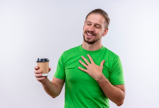 Giovane uomo bello in t-shirt verde che tiene tazza di caffè sorridente allegramente in piedi su sfondo bianco