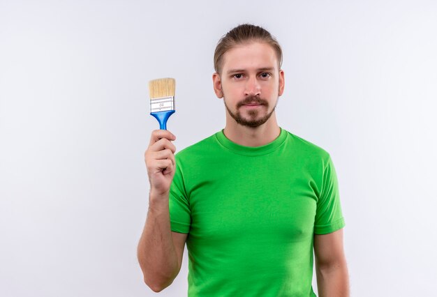 Giovane uomo bello in t-shirt verde tenendo il pennello cercando fiducioso in piedi su sfondo bianco