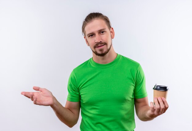 Giovane uomo bello in t-shirt verde tenendo la tazza di caffè guardando fiducioso alzando la mano in piedi su sfondo bianco