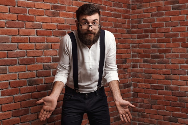 Giovane uomo bello in vetri che gesturing sul muro di mattoni.