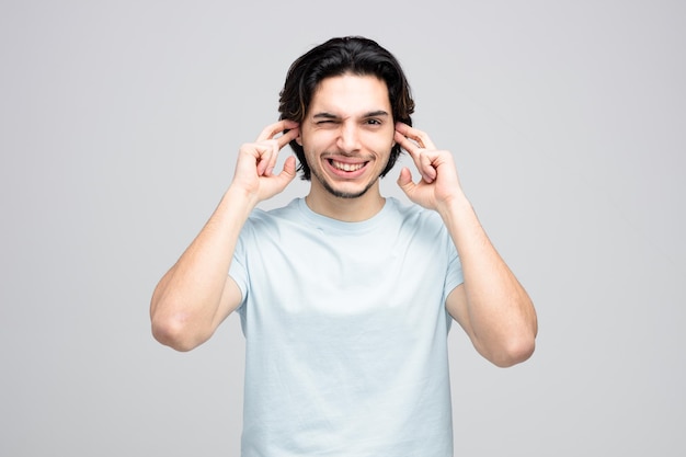 Giovane uomo bello irritato che guarda l'obbiettivo con un occhio chiuso mettendo le dita nelle orecchie isolate su sfondo bianco