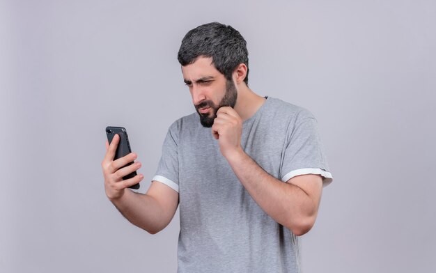 Giovane uomo bello premuroso che tiene e che esamina il telefono cellulare e che tocca il suo mento isolato sulla parete bianca