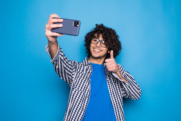 Giovane uomo bello riccio che fa un selfie sopra la parete blu isolata