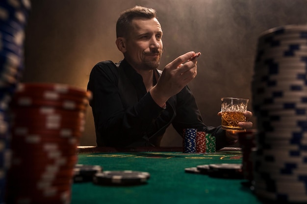 Giovane uomo bello seduto dietro il tavolo da poker con carte e fiches. In primo piano pile di gettoni sul tavolo da poker in una stanza buia piena di fumo di sigaretta.