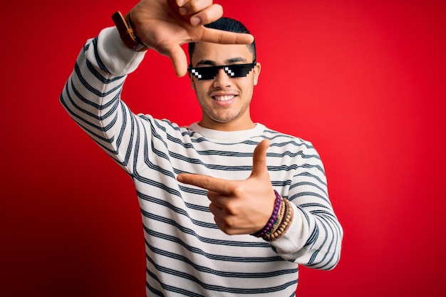 Giovane uomo brasiliano che indossa divertenti occhiali da sole thug life su sfondo rosso isolato sorridente facendo cornice con mani e dita con faccia felice Creatività e concetto di fotografia