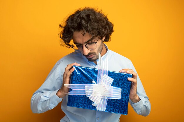 Giovane uomo caucasico bello con gli occhiali tenendo e guardando il pacchetto regalo isolato sulla parete arancione