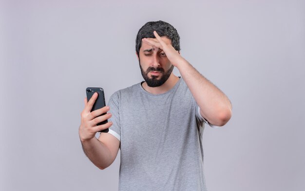 Giovane uomo caucasico bello dispiaciuto che tiene e che esamina il telefono cellulare con la mano sulla fronte isolata su fondo bianco con lo spazio della copia