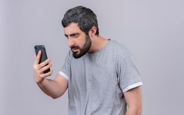 Giovane uomo caucasico bello dispiaciuto che tiene e che esamina telefono cellulare isolato su fondo bianco con lo spazio della copia