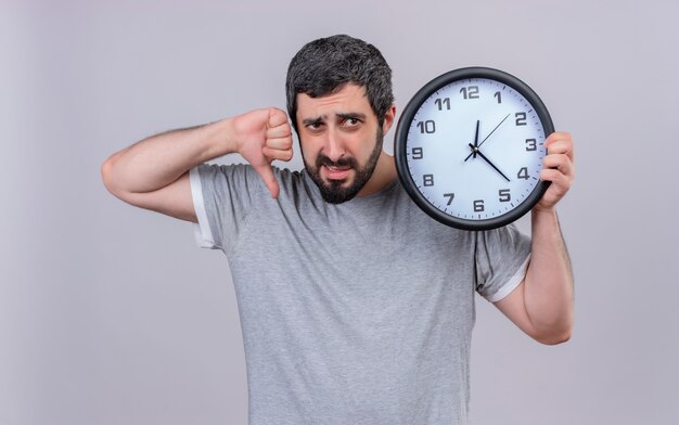 Giovane uomo caucasico bello dispiaciuto che tiene l'orologio rivolto verso il basso e guardando il lato isolato su priorità bassa bianca