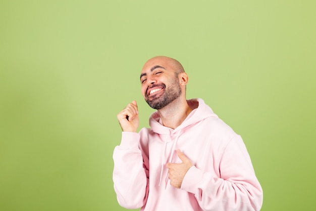 Giovane uomo caucasico calvo in felpa con cappuccio rosa isolato, felice e allegro, sorridente in movimento casuale e fiducioso ascolto di musica