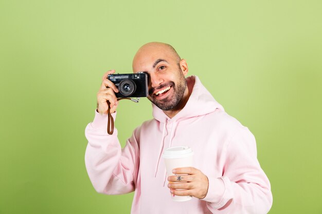 Giovane uomo caucasico calvo in felpa con cappuccio rosa isolato, fotocamera tenuta positiva e tazza di caffè