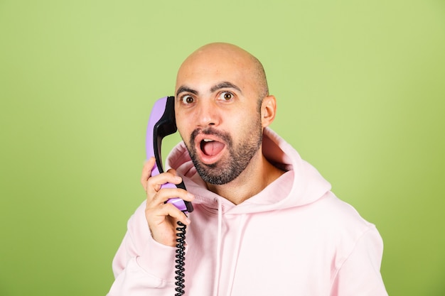 Giovane uomo caucasico calvo in felpa con cappuccio rosa isolato, tenere premuto il telefono di rete fissa con felice faccia stupita eccitata