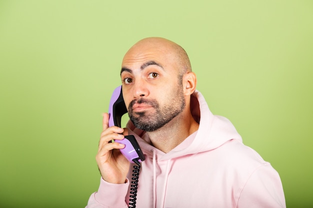 Giovane uomo caucasico calvo in felpa con cappuccio rosa isolato, tenere premuto il telefono di rete fissa con la faccia triste annoiata