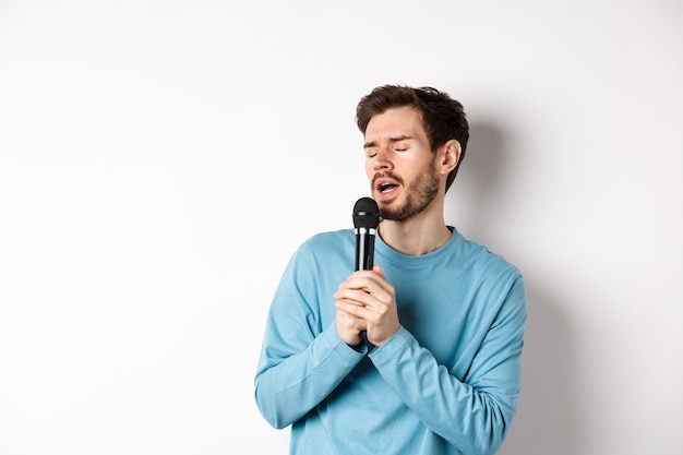 Giovane uomo caucasico che canta una canzone nel microfono con la faccia spensierata, in piedi al karaoke su sfondo bianco.