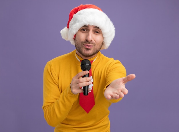 Giovane uomo caucasico che indossa il cappello di Natale e cravatta tenendo il microfono guardando la telecamera cantando allungando la mano verso la telecamera isolata su sfondo viola